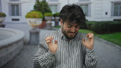 Young man with beard makes a gesture of disgust outdoors on a street with greenery and...