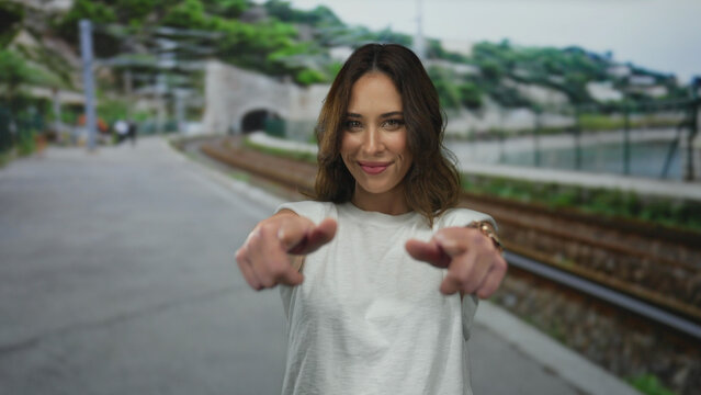 Woman smiling at the outdoor train station, pointing playfully while standing by the railway, with lush greenery and a tunnel in the background.