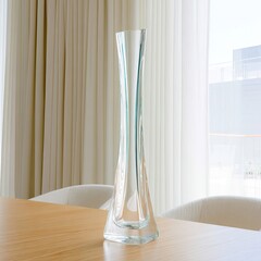 Clear glass vase, tall and slender, sits on a light wooden table in front of sheer white curtains and a window