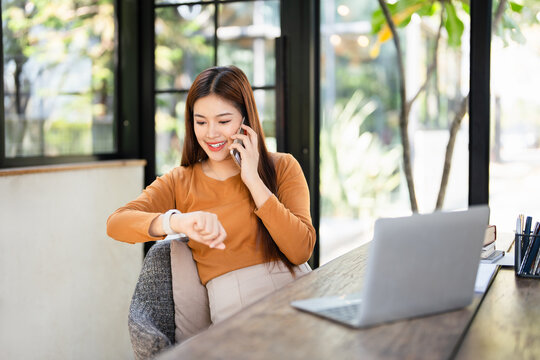 Smiling young asian businesswoman talking on smartphone, checking time on wristwatch. Happy woman arranging meeting with business partners or clients at home office. - Powered by Adobe