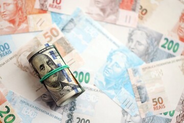 Roll of US dollar bills lies on big amount of Brazilian reais banknotes close up