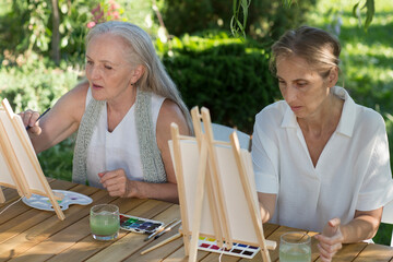 Two happy middle aged woman smiling while drawing watercolor as a recreational activity or therapy outdoors together with the group of retired women. KnowledgeForAll
