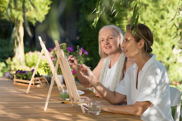 Two happy middle aged woman smiling while drawing watercolor as a recreational activity or therapy outdoors together with the group of retired women. KnowledgeForAll
