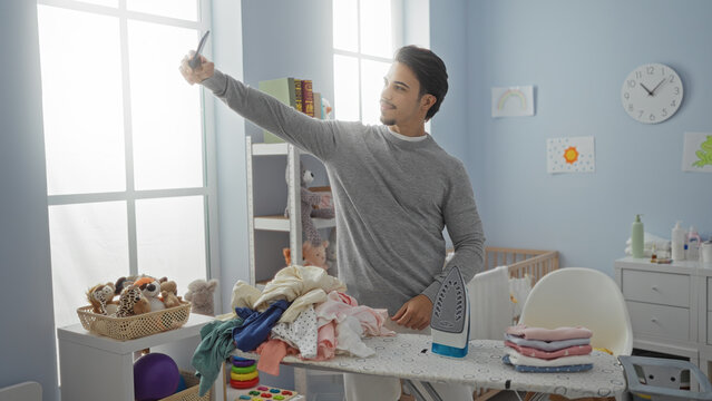 Man taking selfie in laundry room with clothes and ironing board, surrounded by toys and home decor, emphasizing indoor domestic life and personal grooming. - Powered by Adobe