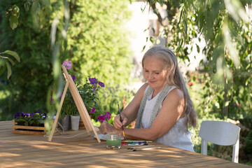 elderly woman with long grey hair paints watercolors sitting at a table in a summer garden. art therapy. online lessons. Dopamine hobbies