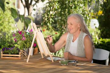 elderly woman with long grey hair paints watercolors sitting at a table in a summer garden. art therapy. online lessons. Dopamine hobbies