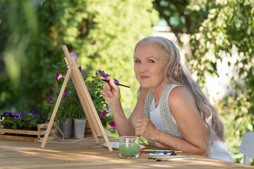 elderly woman with long grey hair paints watercolors sitting at a table in a summer garden. art therapy. online lessons. Dopamine hobbies