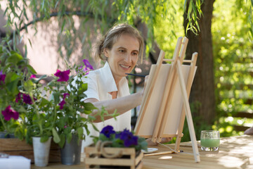 middle aged woman  paints watercolors sitting at a table in a summer garden. art therapy. online lessons. KnowledgeForAll.