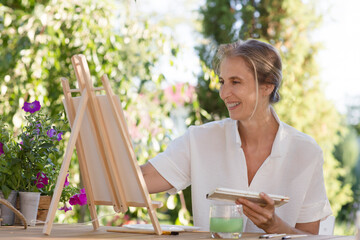 middle aged woman  paints watercolors sitting at a table in a summer garden. art therapy. online lessons. KnowledgeForAll.