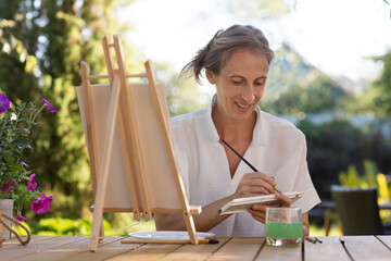 middle aged woman  paints watercolors sitting at a table in a summer garden. art therapy. online lessons. KnowledgeForAll.