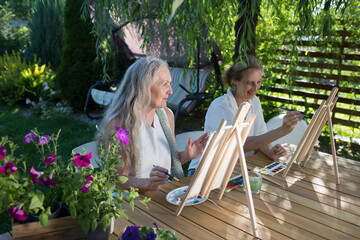Two happy middle aged woman smiling while drawing watercolor as a recreational activity or therapy outdoors together with the group of retired women. KnowledgeForAll
