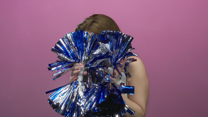 Young cheerleader frowning holds metallic blue pom poms in studio wearing cheerleading uniform;...