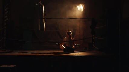 Slender woman enjoying solitude and calmness on boxing ring in gym at night. Athletic lady in sportswear and boxing gloves sitting in lotus position and meditating, recovery body energy and vitality - Powered by Adobe