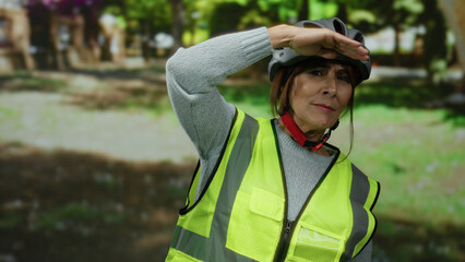 Naklejka premium Senior hispanic woman wearing helmet and reflective vest looks into distance in park setting with trees and greenery surrounding her, embodying outdoor safety and elder activity.