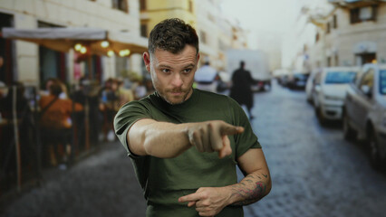 Obraz premium Man in green shirt with tattoos pointing on lively urban street with blurred people and vehicles in background depicting focus and interaction outdoors.