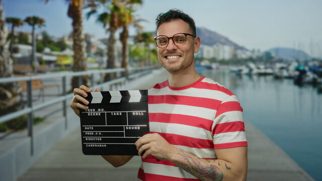 Young man smiling confidently holding clapperboard at vibrant seaside port with boats and palm trees in background under sunny sky.