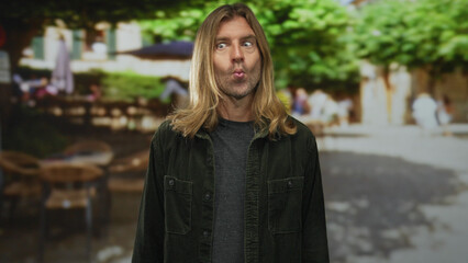 Blond man with long hair in green corduroy jacket puckers lips while looking sideways on a sunny street; curiosity.