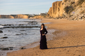 beautiful young woman dancing flamenco walks along the shore of the beach at sunset, wearing a purple velvet dress with ruffles.