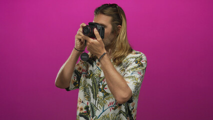 Man wearing tropical shirt holding camera to eye in bright pink studio; creativity exploration...