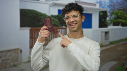 Smiling man in front of white building holds passport and points finger to passport; travel...