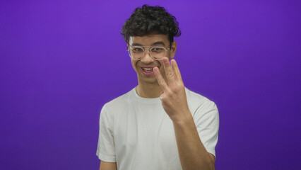 Young man wearing glasses and white t shirt holds up three fingers in purple studio; celebration...