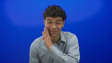 Young hispanic man cups hand to cheek while whispering in studio with blue background; secrecy...