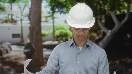 Young hispanic man wearing hardhat reviews blueprints while smiling near building; planning...
