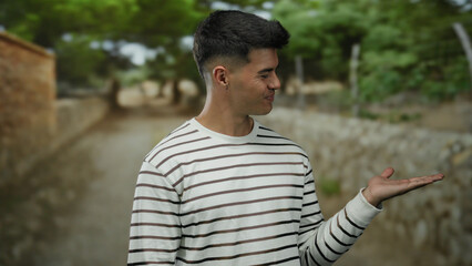 Young man with striped shirt and open hand standing outdoors in a park setting with a stone path...