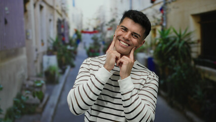 Young man smiling confidently on a vibrant urban street wearing a striped shirt, surrounded by lush...