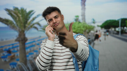 Hispanic man with backpack gives thumbs up while talking on phone at seaside promenade outdoors...
