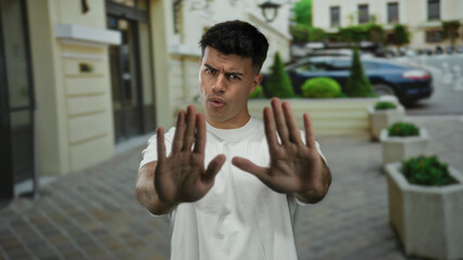 Fototapeta premium Man standing on city street with hands raised as if signaling stop wearing a white shirt in an urban setting on a sunny day showcasing expressive body language and serious expression.