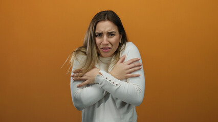 Young blonde woman feeling cold in a white sweater, hugging herself against an isolated orange background, expressing discomfort and shivering slightly.