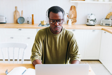 Young black man with headphones and glasses concentrating on working on his laptop in a modern,...
