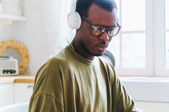 Professional african american male focusing on laptop screen, wearing noise-canceling headphones and stylish eyewear in minimalist home workspace, representing contemporary remote work environment