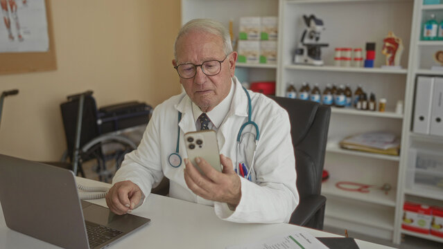 Senior male doctor in clinic examines smartphone near laptop in medical office. - Powered by Adobe