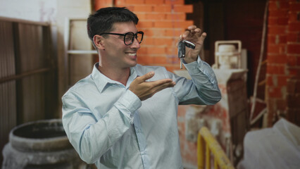 Young man holds key fob between fingers in building with exposed brick interior and scaffold beams;...