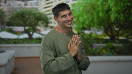 Man in green sweater clasping hands at chest on street next to building facade; serenity gratitude moment.