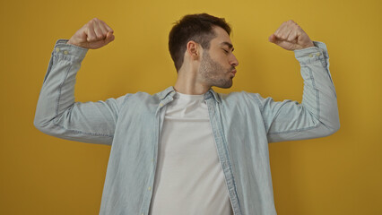 Young hispanic man posing confidently with arms flexed against isolated yellow background in a...