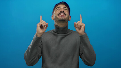 Man in gray turtleneck smiling and pointing both index fingers upward in blue studio setting;...