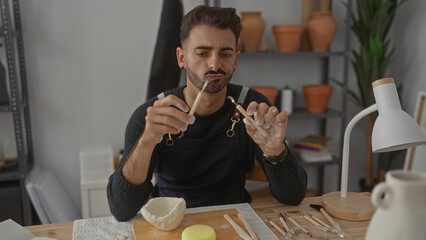 Man holds carving tool with clay covered hands at wooden worktable in studio; concentration...