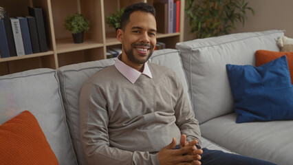 Young man sitting relaxed on a comfortable sofa in a modern living room, smiling warmly, surrounded...