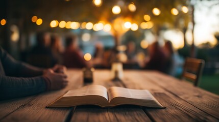 Open bible laid flat on the table while group of people enjoying the outdoors at sunset with the string light in the blurred background