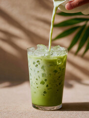 Creamy milk being poured into a glass of iced matcha latte with fresh green leaves in soft natural light setting