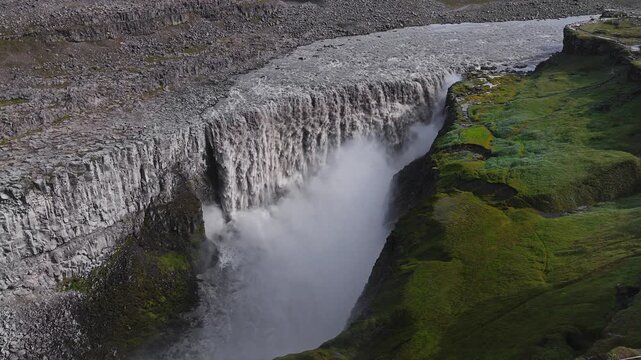 Dettifoss waterfall in Iceland plunges into a canyon with mist rising, rugged terrain, and green moss. The water's motion is dynamic and forceful.