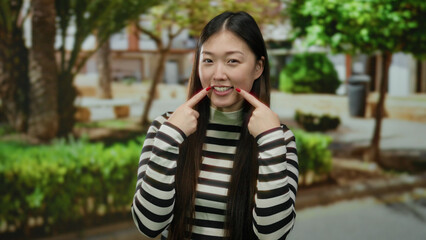 Woman smiling in a striped shirt standing on a sunny urban street with trees in the background,...