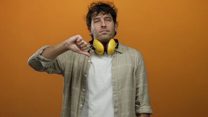 Hispanic man wearing yellow noise-canceling headphones showing thumbs down against an orange...