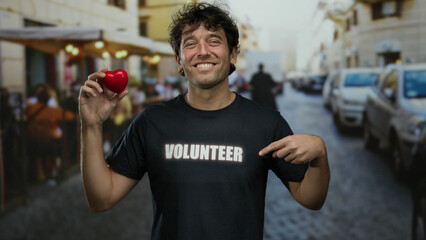 Hispanic man smiling outdoors in a street while holding a red heart and wearing a volunteer shirt, depicting charity and community spirit.