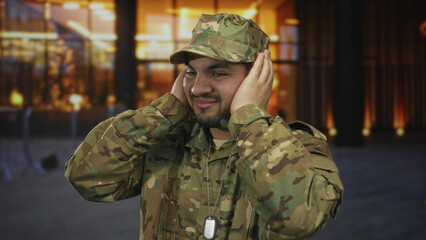 Young hispanic soldier in green camouflage uniform covers ears with bare hands on a city street;...