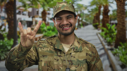 Man in camouflage uniform displays dogtag and rock horns gesture in green park; fun metal...