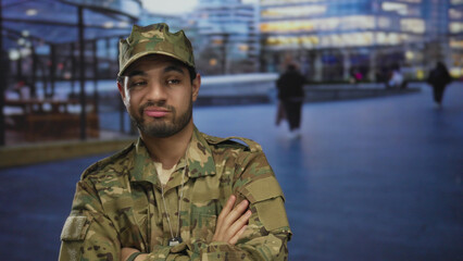Hispanic soldier in camouflage uniform with dog tags stands with arms crossed on a city street;...